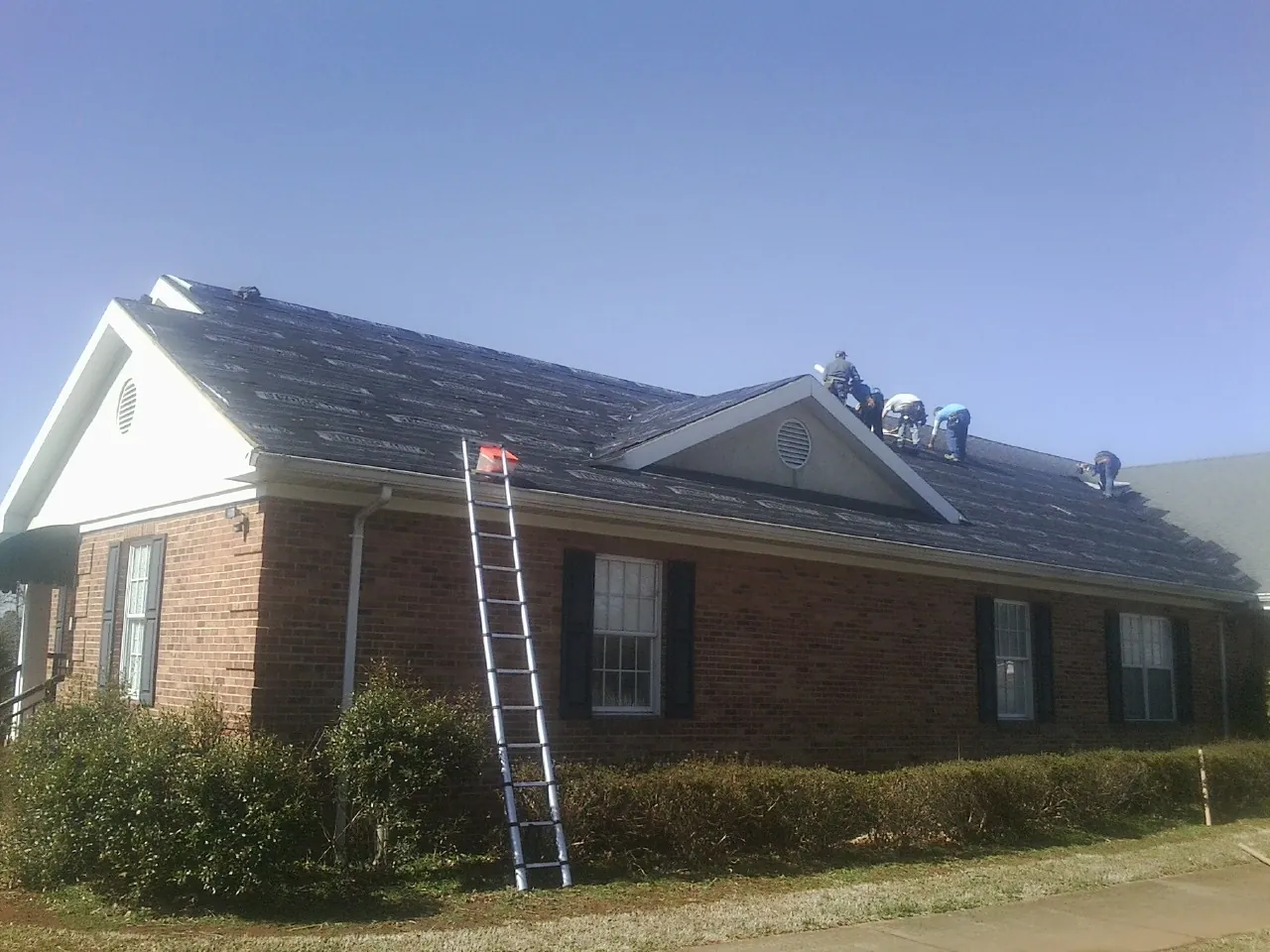 Crew tearing shingles off a brick ranch roof with a ladder set — Emergency Roof Repair work by our crew in Pilot Point