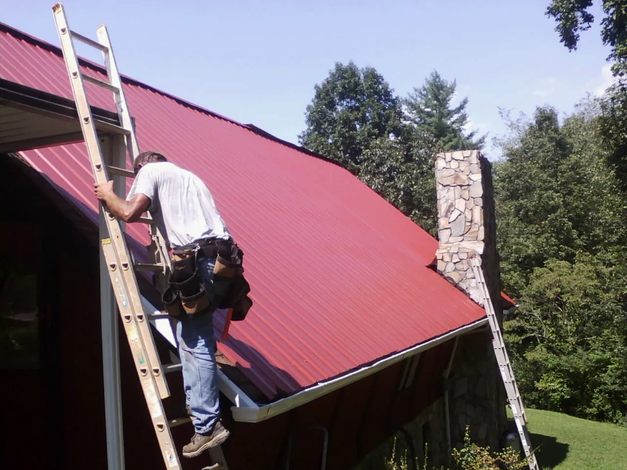 Roofer on a ladder beside a red metal roof and stone chimney — Soffit & Fascia Repair by Roofing Brothers in Pilot Point